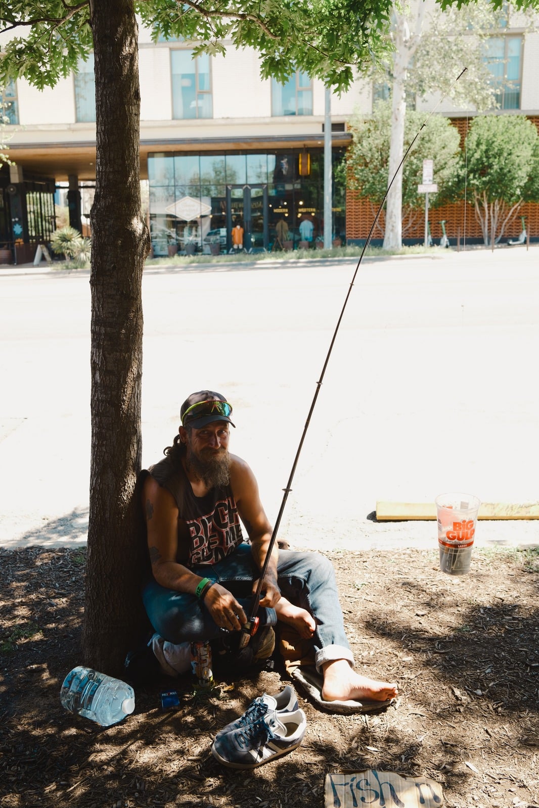 A bearded man sits barefoot under a tree beside the street, holding a fishing rod; a cardboard sign reads “fish’n”