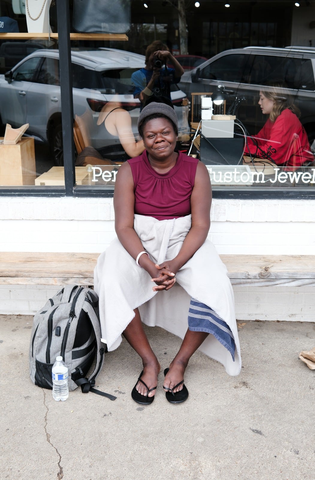 A woman sits on a bench outside a jewelry shop, a backpack and water bottle at her feet; the photographer is reflected in the window