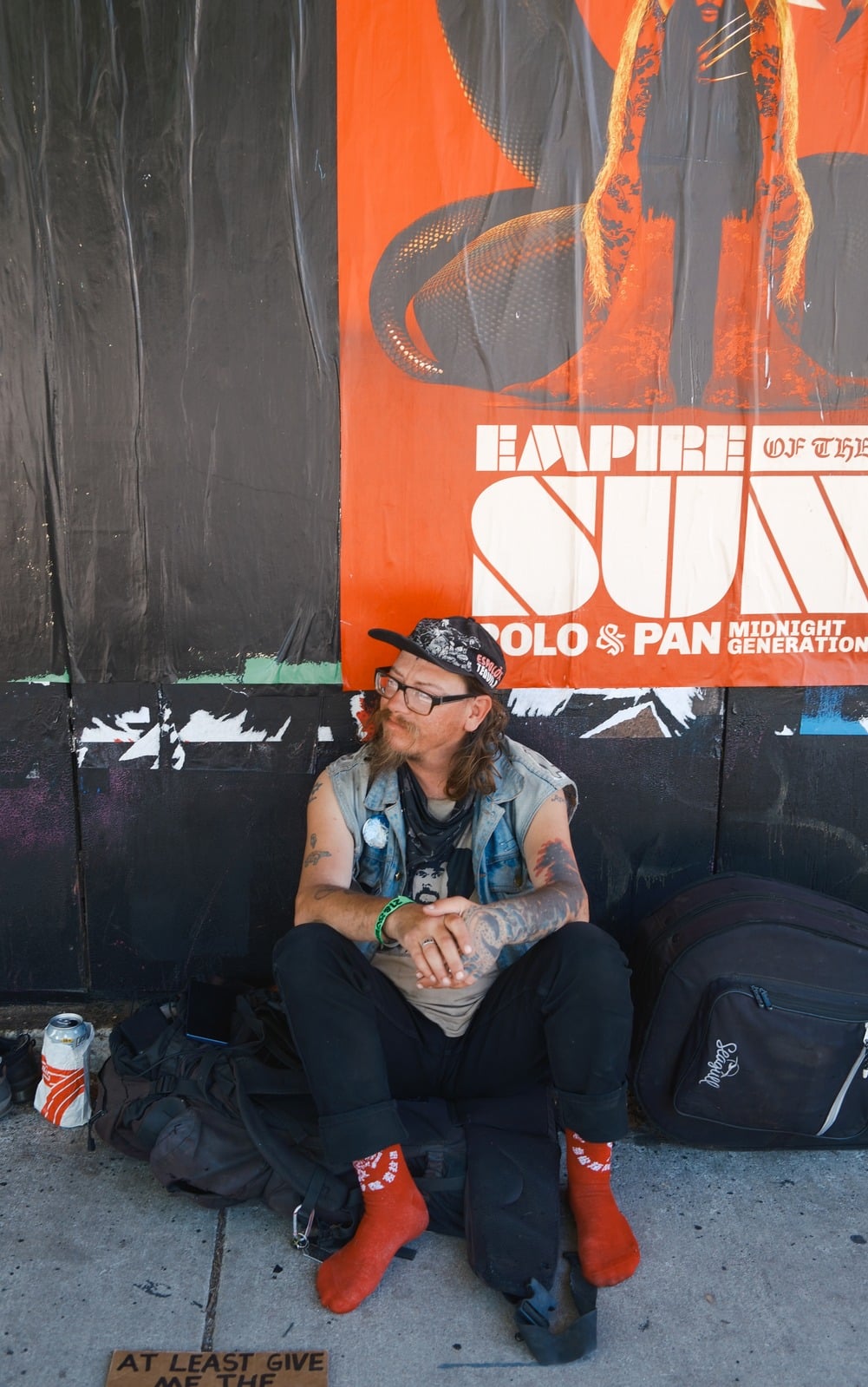 A man in a cap and glasses sits on the sidewalk beneath an orange concert poster