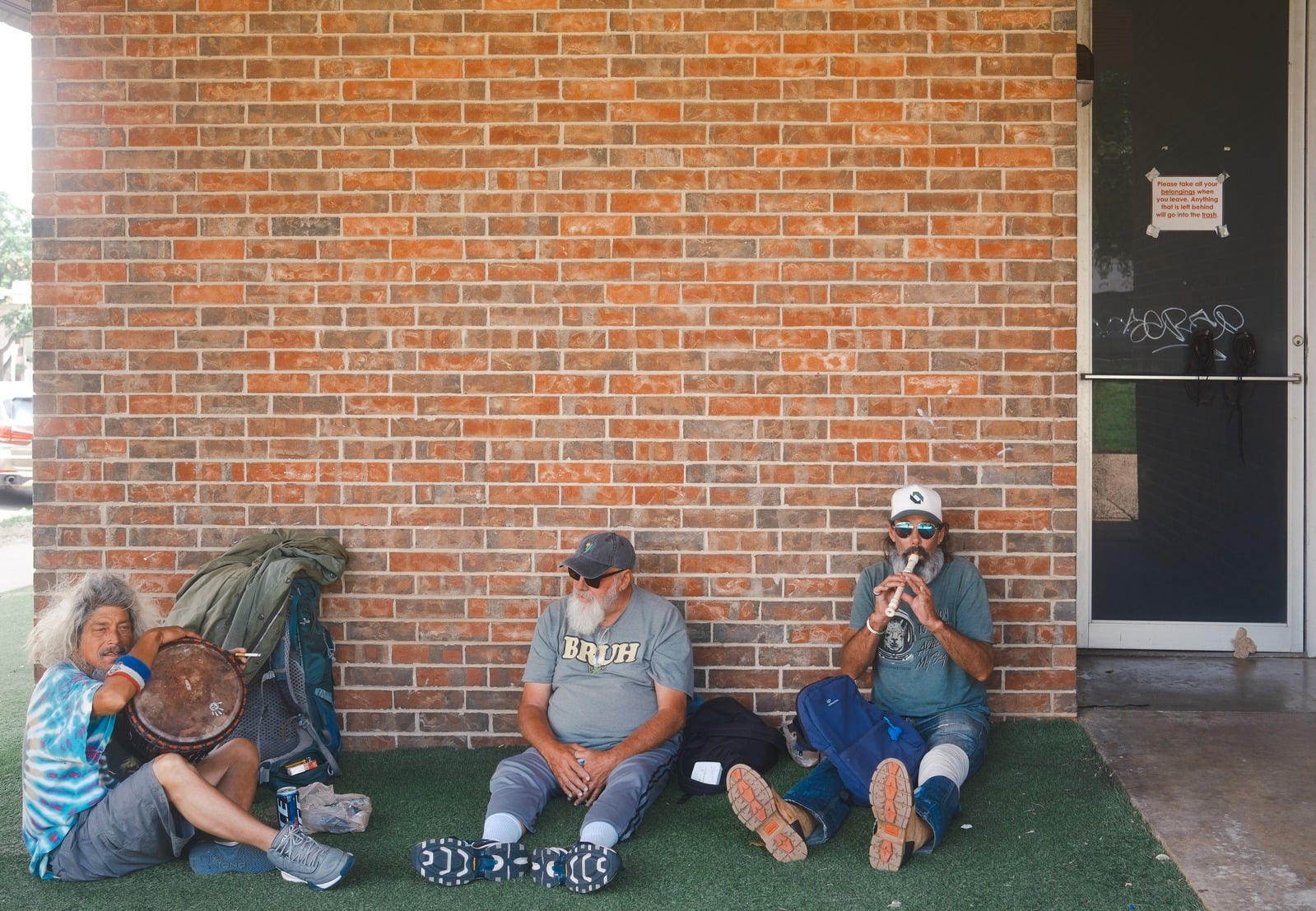 Three men sitting against a brick wall; one plays a recorder, another holds a hand drum