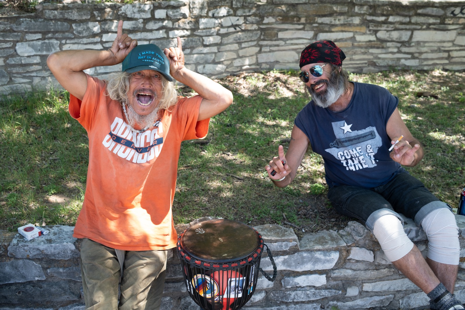 Two men laughing on a stone wall in a park, a hand drum between them